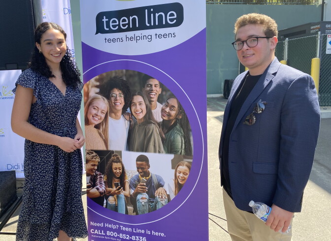 Teen Line counselors Lily Kramon (L) and Jonathan Gelfond (R) completed 65 hours of training and months of shadowing in order to support their peers over calls and texts. They stand on either side of a banner that features pictures of youth hanging out and smiling. The banner reads "teen line, teens helping teens." 