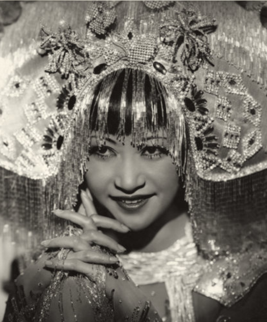 Anna May Wong in her role as Princess Ling Moy. She wears an elaborate headdress that creates a half circle atop her heard, embroidered with beaded filigree. Sparkly fringe falls from the bottom of the headdress framing Wong's face with silver bangs. She smiles looking up from hooded eyes, seemingly micheviously. 