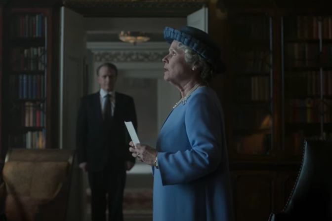 A profile of woman in a blue dress and blue hat holding a piece of paper as a man in a black suit in the distance looks at her. Book shelves line the walls.