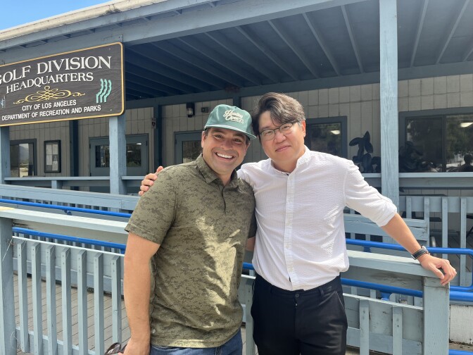 Two men, one in a ball cap, embrace side-by-side and smile for a photo.