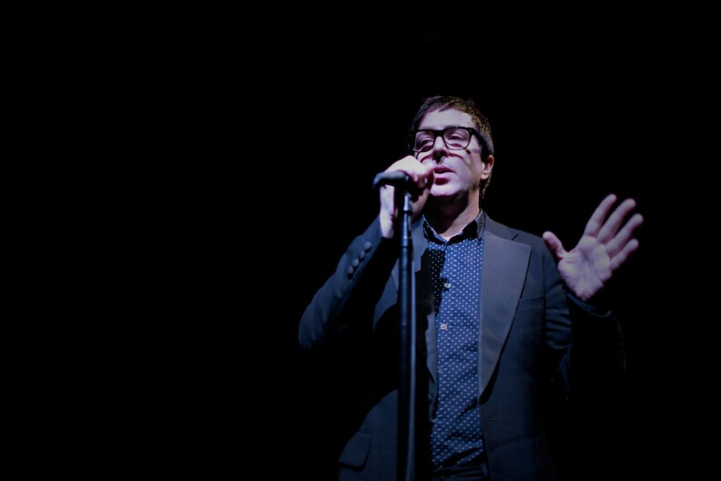 a man in a dark suit, polka dotted blue button down shirt and glasses stands at a microphone and talks