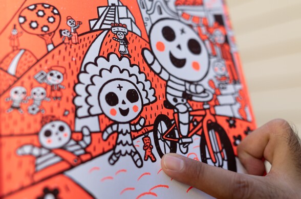 A man's finger points at the illustration of a small orange worry doll that looks a bit like a carrot. The doll has a look of concern. A skeleton girl and a skeleton boy on a bicyle are next to the doll on the sidewalk.
