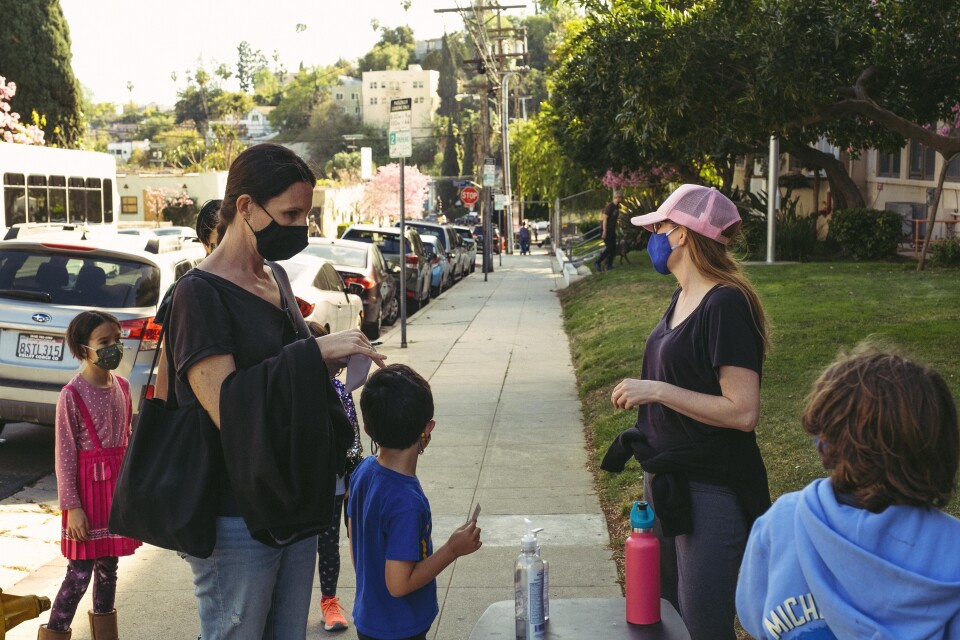 A woman in a pink baseball hat and blue facemask is talking to parents and children on a sidewalk.