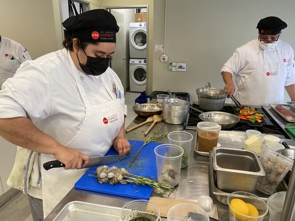 Culinary apprentice Nicole Rios, in a white chef's outfit with a black facemask, stands in a kitchen cutting a piece of rosemary on a blue cutting board next to a pile of shrimp skewered on rosemary twigs while another chef grills vegetables nearby.