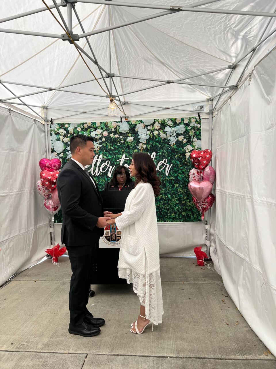 A man wearing a black suit and a woman wearing a white dress stand in front of officiant clutching hands. In the backdrop a green, leafy banner is visible as well as heart shaped balloons on either corner. 