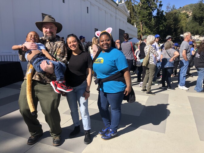 A light-skinned man with a thick goatee wearing a broad hat holds a brown-skinned boy wearing jeans and red sneakers in his arms. Beside him is a woman with light brown skin wearing a black t-shirt and jeans. Beside her is a woman with dark brown skin wearing a headpiece resembling mountain lion ears, a light blue t-shirt, and darker blue jeans.
