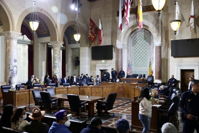 The council chamber dais is empty as people stand and head out for recess. Members of the public in the front row remain seated. Various police officers surround the dais. 