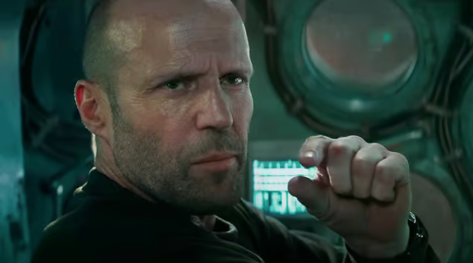 Jason Statham sits in a submarine, the video controls visible behind him. He wears an all back water-suit. He looks off at someone out of frame. His eyes slightly hooded by his eyebrows pressed together, giving a serious look. He holds up one hand his index and thumb in a pinch gesturing to the size of something perhaps. 