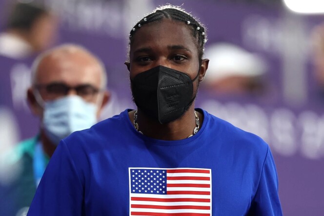 A man has a black mask on his face and is wearing a blue shirt with an American flag in the middle of it 