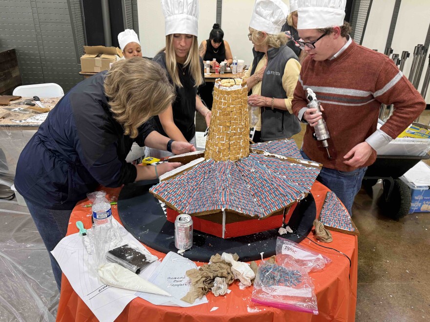 A group of people wearing chef hats are working to build a gingerbread structure