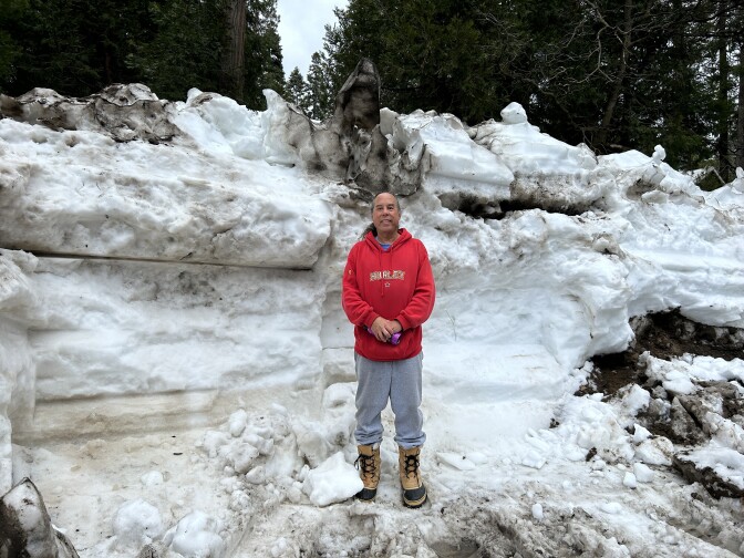 A man in a red sweatshirt and grey sweatpants an snow boots stands in front of a snow berm taller than his head. 