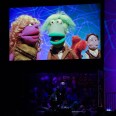 A view of about three puppeteers dressed in black on a stage with a large screen to their left showing a purple female puppet a green male one, a purple male one with glasses in the back and what appears to be a squirrel puppet next to him.
