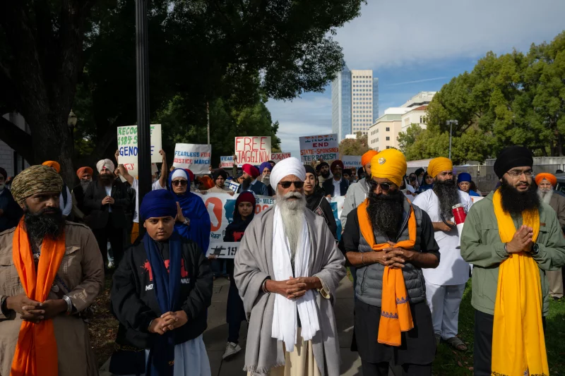A crowd of people wearing traditional garments and turbans are walking together outside carrying signs and holding their hands together