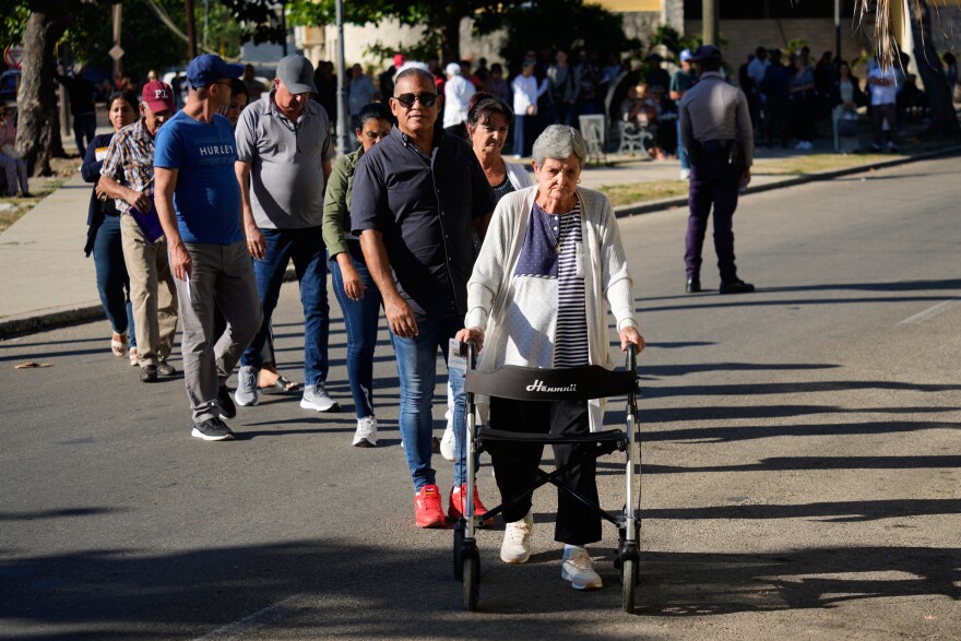 People line up crossing a street. There's a line and crowd of more people in the background.