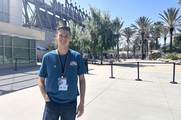 A teenage boy wearing a blue t-shirt and denim jeans is posing with one hand in his pocket. He's smiling at the camera on a sunny, clear day. 