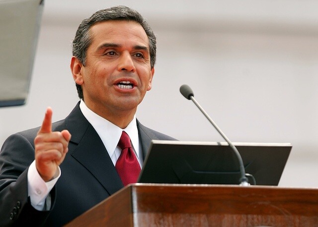 A man an a lectern wears a dark suit jacket, white shirt and red tie
