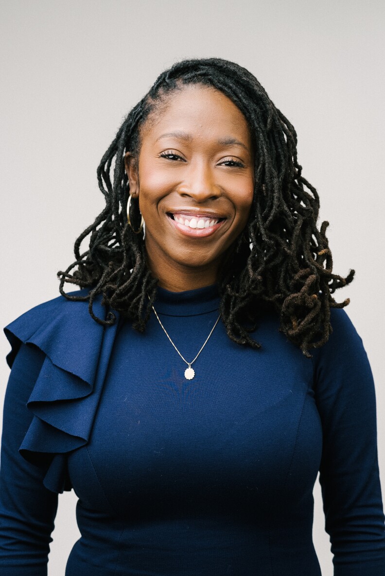 A Black woman with shoulder-length hair wearing a Navy blue long-sleeved shirt smiles for the camera in a headshot portrait.