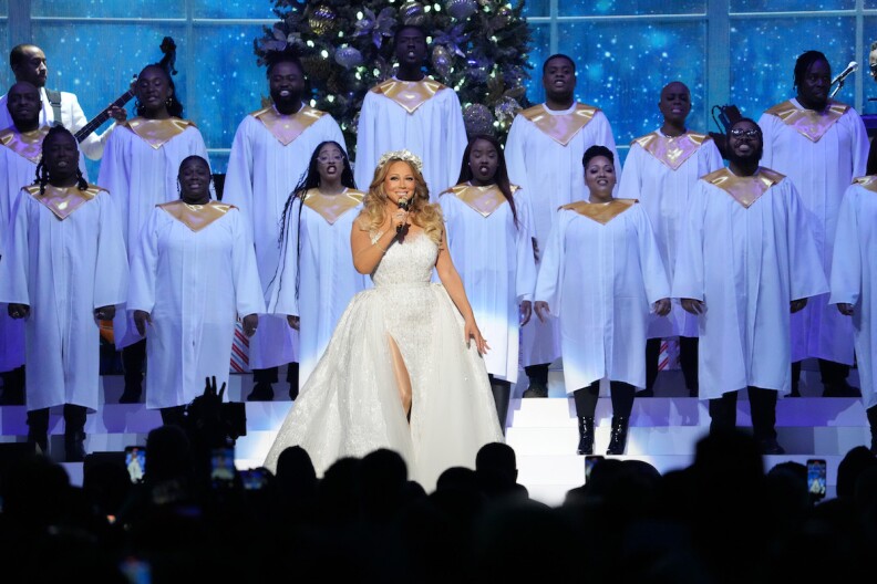 Mariah Carey, dressed in an all-white ballgown, sings on stage with a choir in white robes behind her.