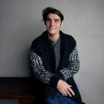 RJ Mitte, a smiling person with dark, tousled hair, sits against a gray studio wall, looking off to the side. They wear a black-and-white patterned knit cardigan over a gray henley.