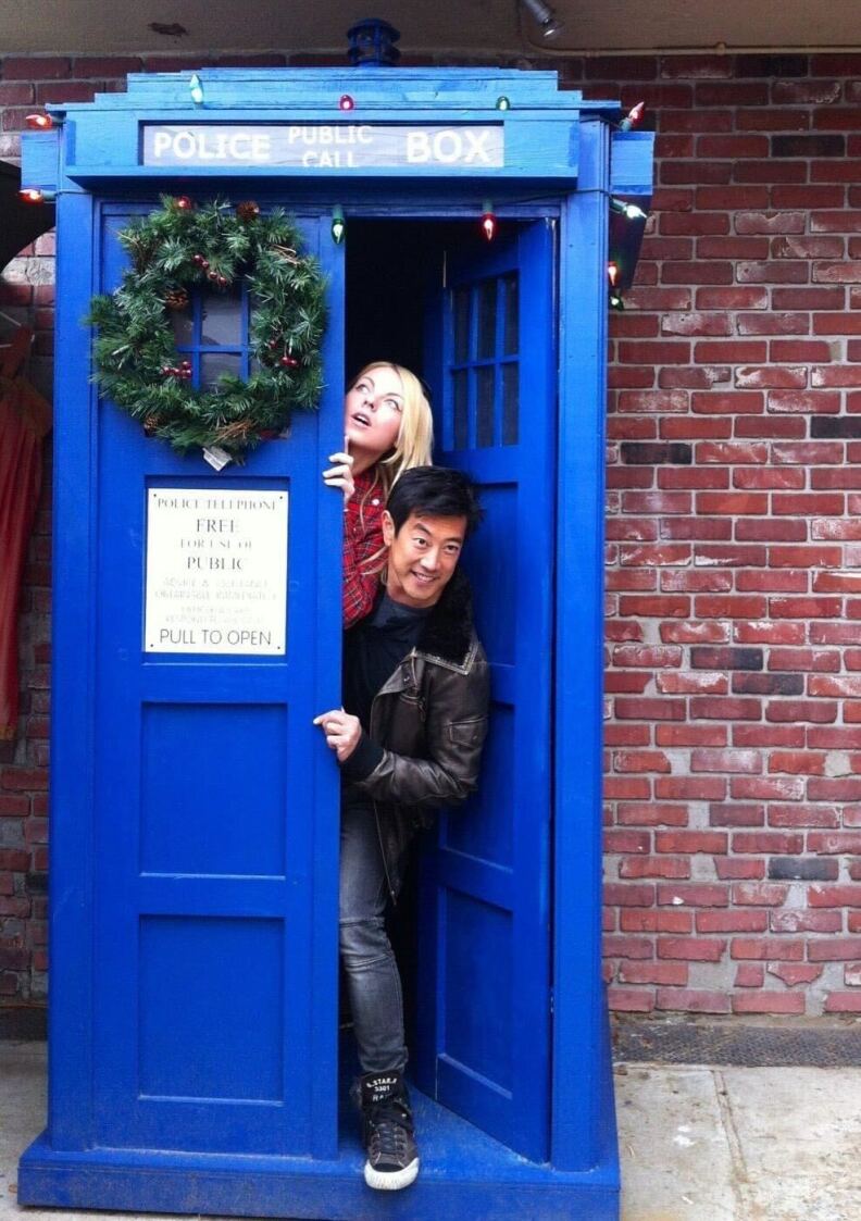 A blue police telephone box in front of a red brick wall, with a man and woman peeking out from behind the door.