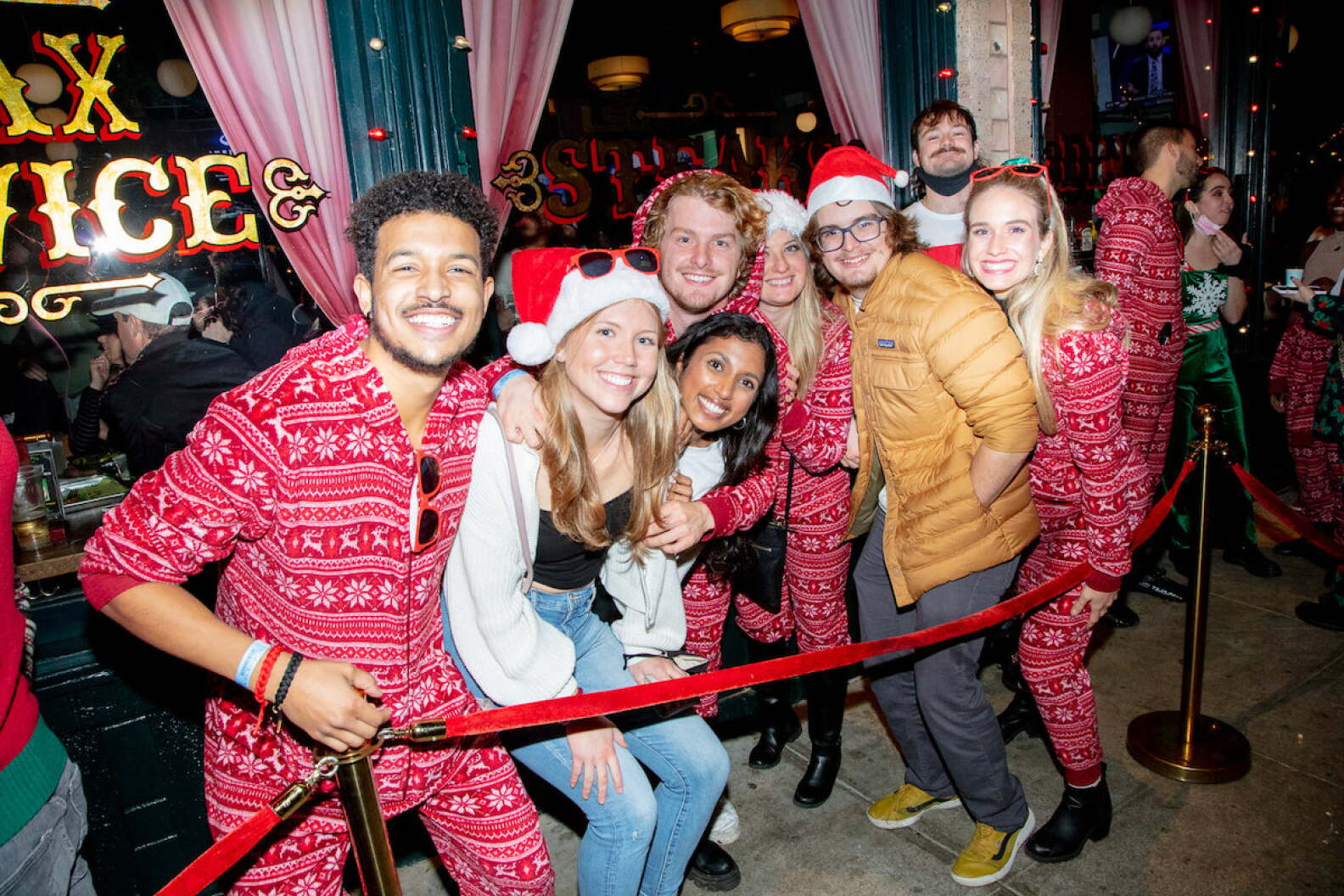 A group of friends wearing holiday pajamas and Santa hats stand in line behind red velvet stantions.