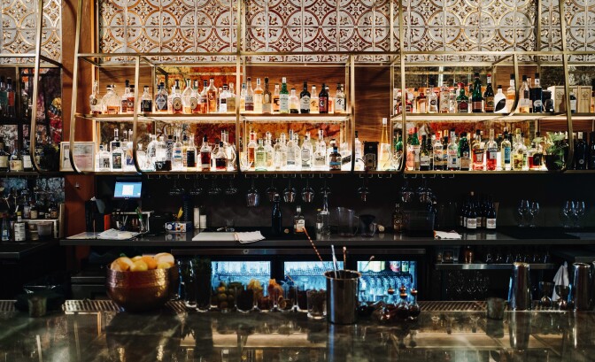 A back-lit bar contains various bottles of alcohol, with rows of cocktail glasses underneath. In front, there's a large metal-topped counter  