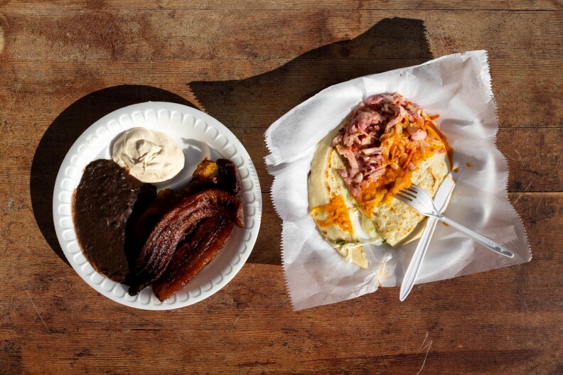 Two side-by-side dishes sit on disposable plates placed on a wood tabletop: On the left, there are soupy black beans, plantains fried to a deep dark brown, and a generous dollop of sour cream. On the right, there's a pupusa that has been cut into, with the cheese oozing out. The pupusa is drizzled with a ribbon of orange salsa. A plastic knife and fork lay nearby, ready for digging in.