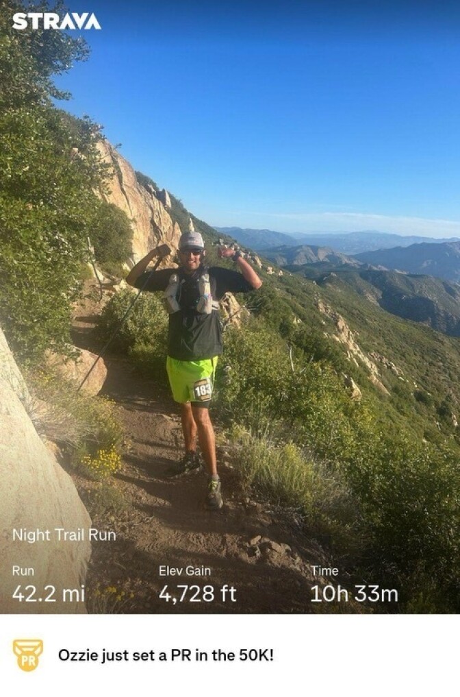 A man poses on a dirt trail in the mountains. He's wearing sunglasses and a hat. 