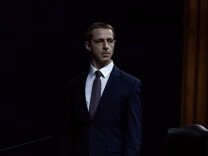 A male-presenting person with a strawberry blonde buzz-cut stands in a dark room wearing a white button-up dress shirt, a gray tie, and a navy blue suit jacket. There is a yellow curtain behind him on his right, and the upper part of a leather chair is visible in the bottom right corner of the image.