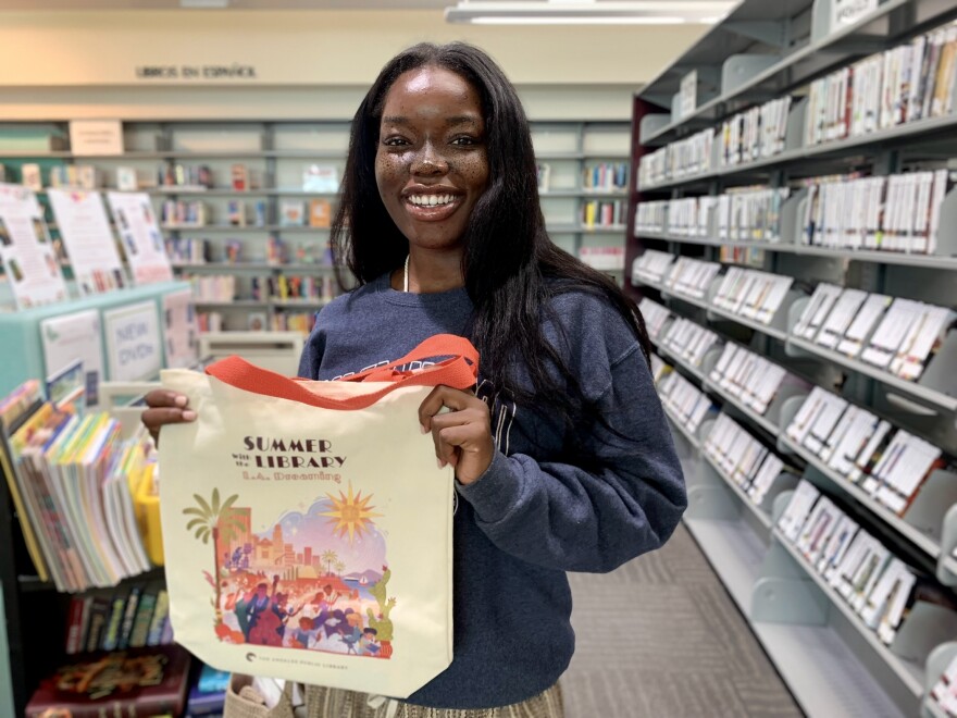 A woman with dark skin tone and long dark brown hair smiles and holds up a cream colored canvas totebag that reads Summer with the Library and has a colorful scene of Los Angeles including a palm tree, sun, and prickly pear cactus. She is surrounded by full bookshelves. 