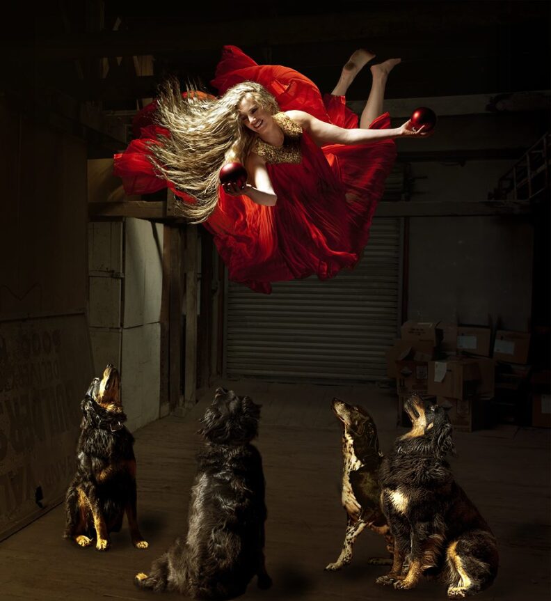 A blonde woman in a red dress floats in the air holding a ball while four dogs underneath her look up.