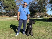 Eddie Aldrete stands on green grass at a park, holding the lede of his dog. The dog is a black Australian Labradoodle named Dakota. 