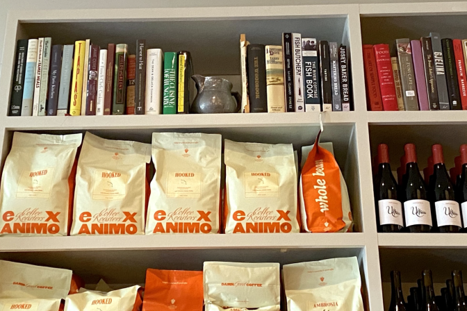 A bookcase has a series of cookbooks on the top shelf, with cream and orange coffee bags and bottles of wine below.