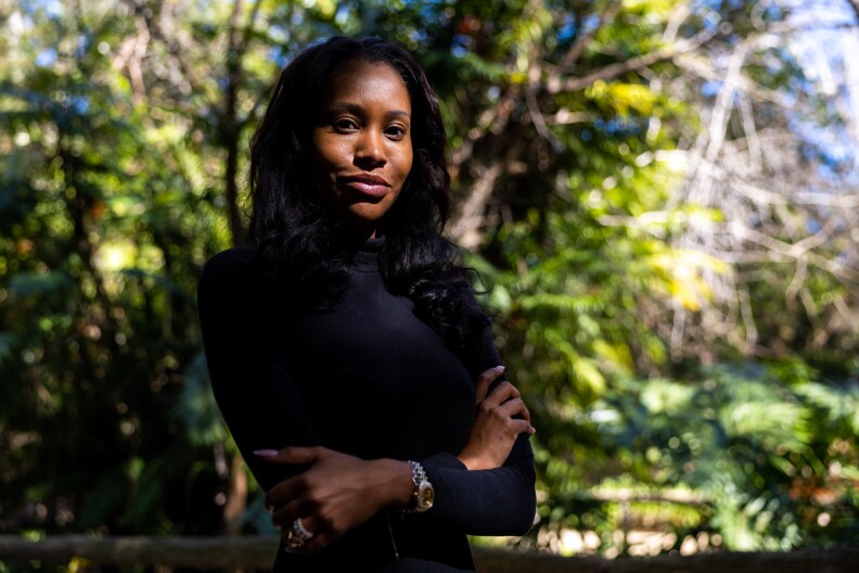 A dark-skinned woman with shoulder length black hair stands with her arms crossed in front of her body. She is wearing a black turtle neck and is standing in front of a group of green trees that are out of focus.