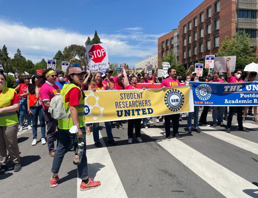 Student protesters, some in hot pink shirts, hold up a banner that read "Student Researchers United," along with signs that read "STOP exploiting workers." The students are in the middle of the street, standing on a crosswalk.