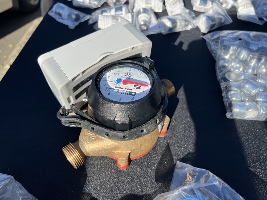 A grey plastic box is attached via a rubber strap to an example of a metal water meter, which has a clock-face device with a hand that points at the latest reading of the amount of water — in cubic feet — that has flowed through the pipe. The device sits on top of a black tablecloth and is surrounded by other various metal hardware.