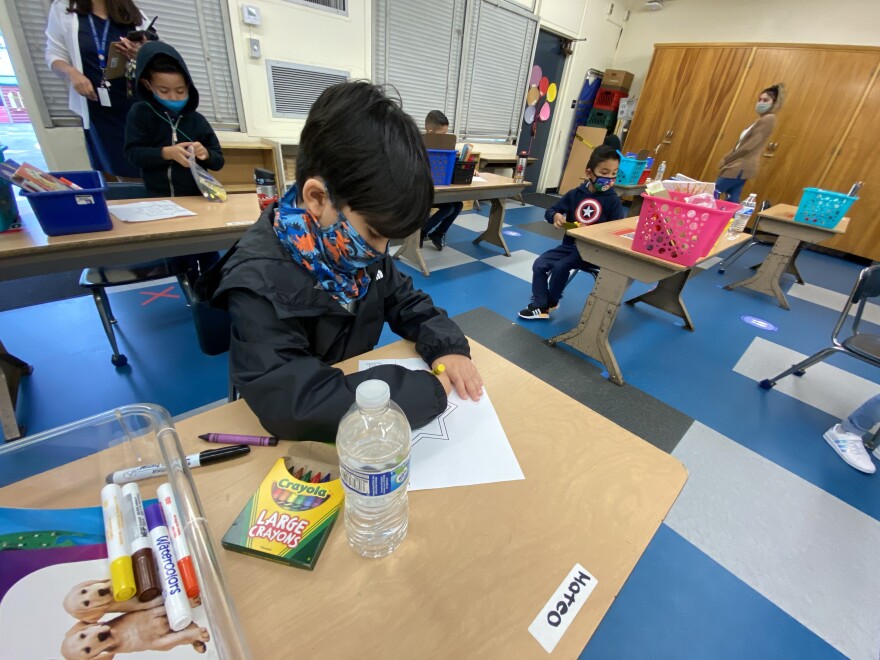 A small child works at his desk with paper and crayons. He wears a blue and orange mask. The desks in the room are spaced several feet apart.