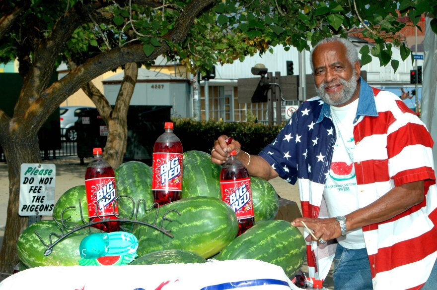 A dark skinned Black man in a shirt decorated as a American flag poses with three Big Red soda bottles and several watermelon.