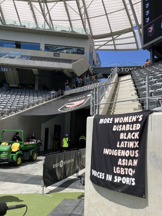 A black fabric sign draped over the edge of a section of BMO Stadium near the field reads "More women's, disabled, Black, Latinx, Indigenous, Asian, LGBTQ voices in sports" in pink letters. Visible in the background is the Angel City Football Club logo and empty stadium seats.