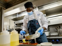 A man in a dark blue apron and light-blue medical mask places food from a frying pan into a pan. Behind him is a large industrial stove.