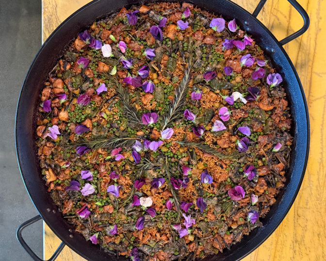 An overhead shot of a large black paella pan filled with saffron-orange rice studded with chicken, charred green vegetables, and green peas, arranged in a starburst pattern with sprigs of rosemary and scattered purple and pink flower petals. The pan sits on a mustard-yellow tabletop.
