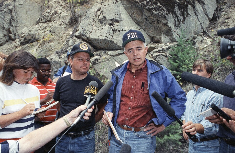 A man with light skin tone, wearing an FBI hat, blue jacket, and red shirt, speaks in front of reporters holding microphones and some writing notes. Behind them is a mountain side with large boulders and moss growing on them.