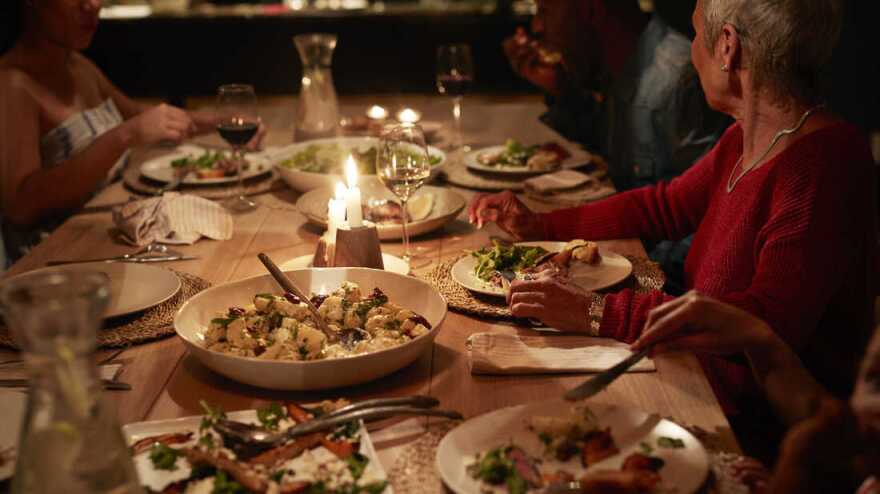 A wood table has food served family style including potatoes and salad with glasses of red and white wine. People sitting at the table have medium-skin tones.
