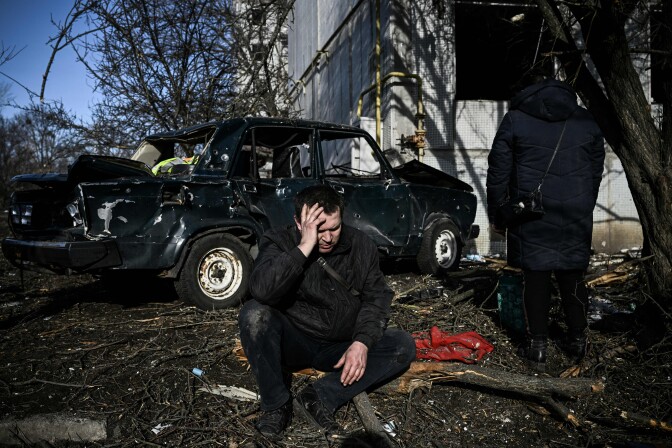 A man kneels and holds his head as he looks down with a distraught expression on his face. A severely damaged car is behind him, with sticks and other dirt surrounding him. Another person has their back to the camera and wears a large Black jacket. 