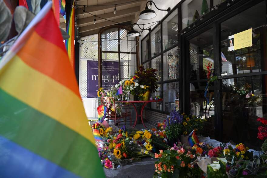 A rainbow flag hangs in the foreground. In the background, there are bouquets of colorful flowers stacked outside of a clothing store called Mag.Pi.