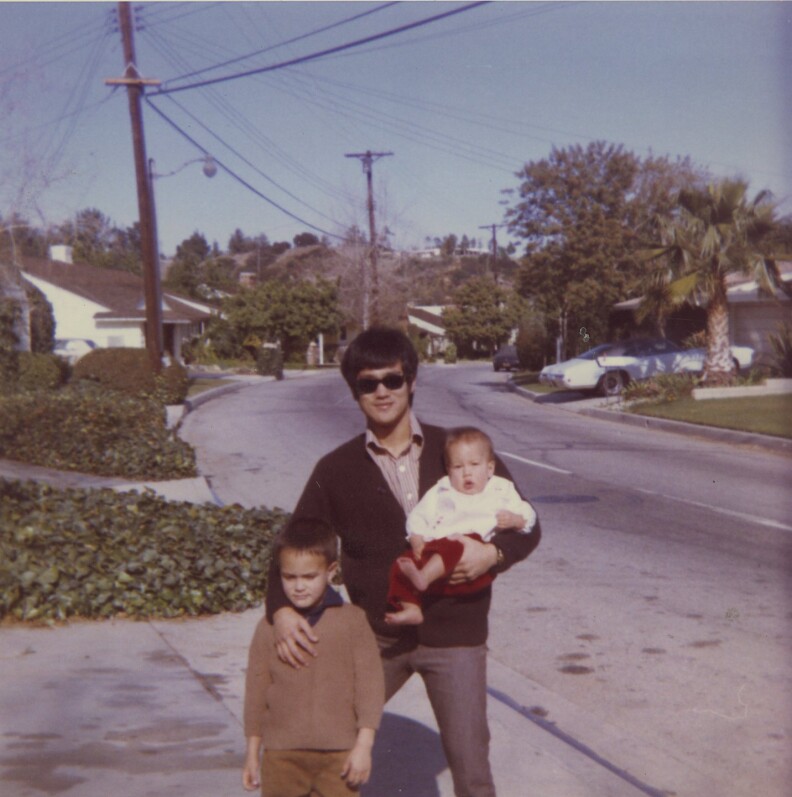 An Asian man in a dark sweater, brown shirt and sunglasses smiles in driveway of home holding his baby daughter, while resting with hand on his son's chest.