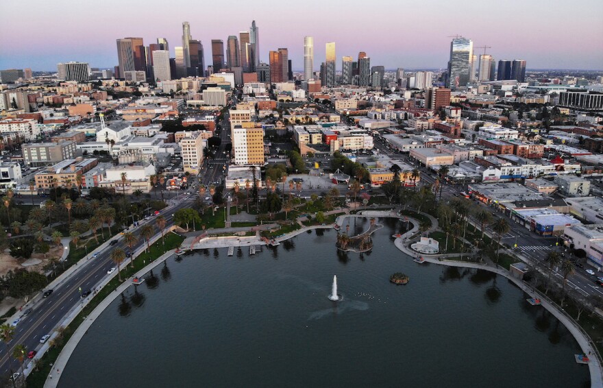 An aerial view of the city of Los Angeles with the lake at MacArthur Park in the foreground. In the distance is the Downtown Los Angeles skyline with a row of tall office buildings at the center of the photo.