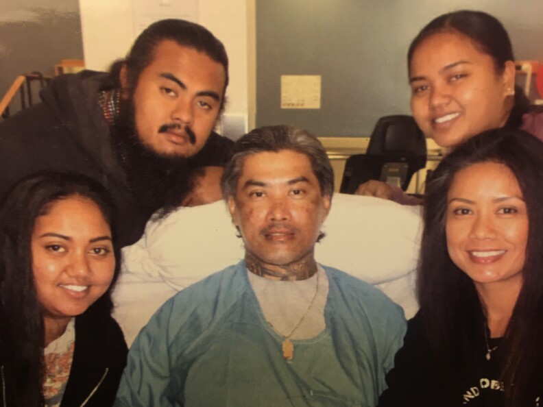 A Cambodian man in a hospital bed is surrounded by four smiling relatives-- a man and woman on his left, and two women on his right.