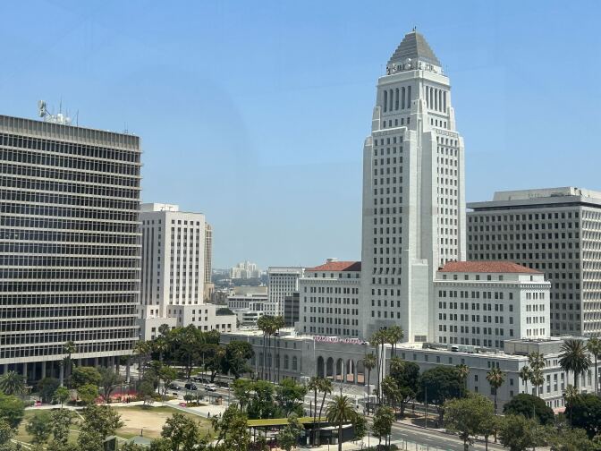 A tall, white building is surrounded by shorter buildings and trees during the day.