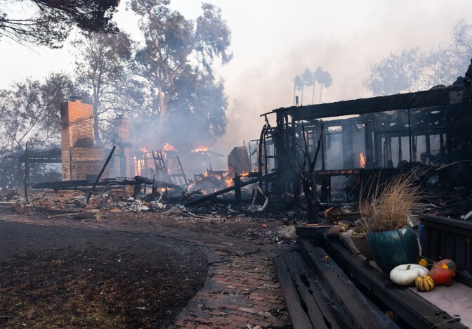 A burnt down property. On the far right of frame some planters and pumpkins rest amidst the damage. Fire can be seen burning in the background. 